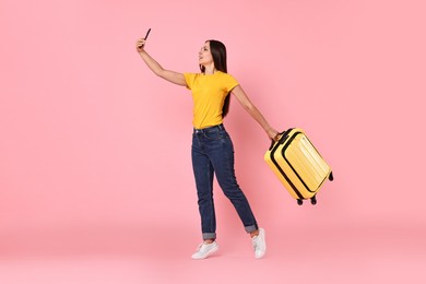 Photo of Happy young woman with suitcase taking selfie on pink background