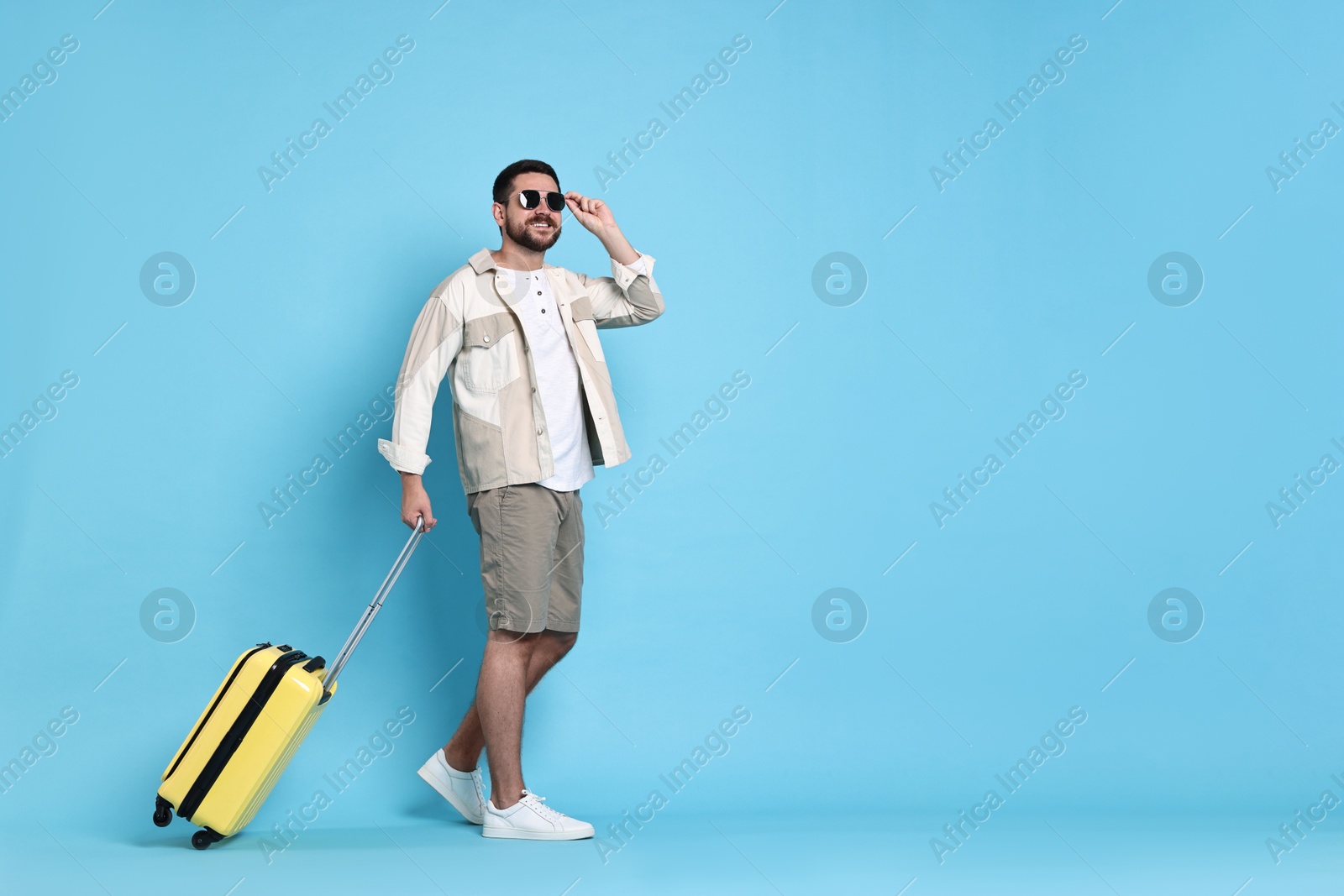 Photo of Happy man in sunglasses with suitcase on light blue background, space for text