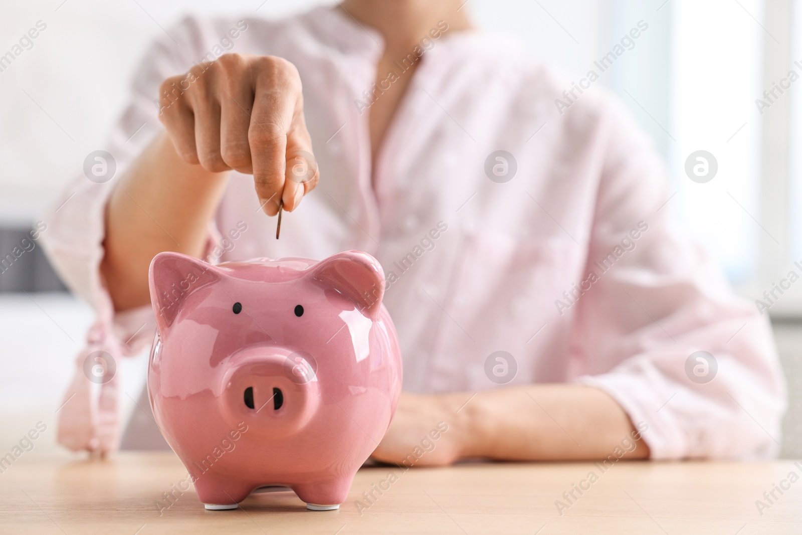 Photo of Woman putting coin into piggy bank at table indoors, closeup