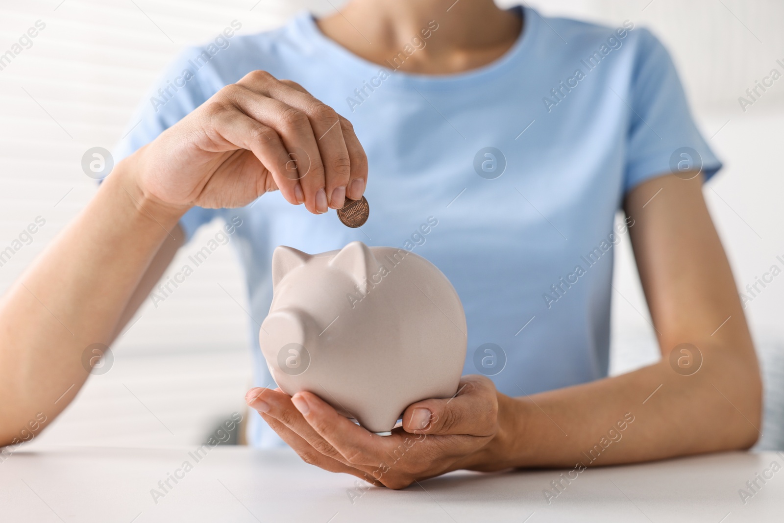 Photo of Woman putting coin into piggy bank at white table indoors, closeup