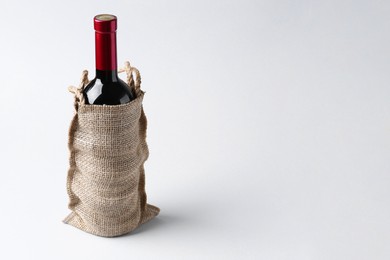 Photo of Wine bottle in burlap bag on light grey background, space for text