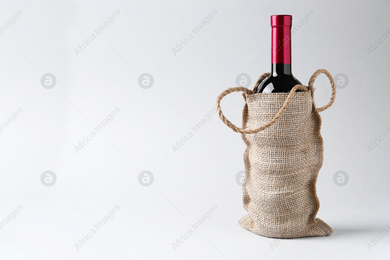 Photo of Wine bottle in burlap bag on light grey background, space for text