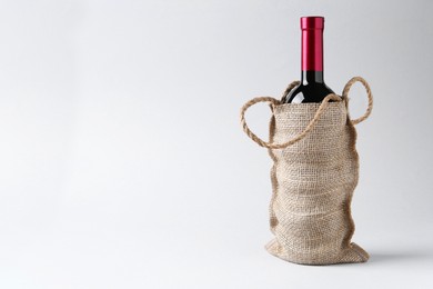 Wine bottle in burlap bag on light grey background, space for text