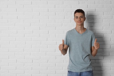 Photo of Teenage boy wearing grey t-shirt and showing thumbs up near white brick wall, space for text