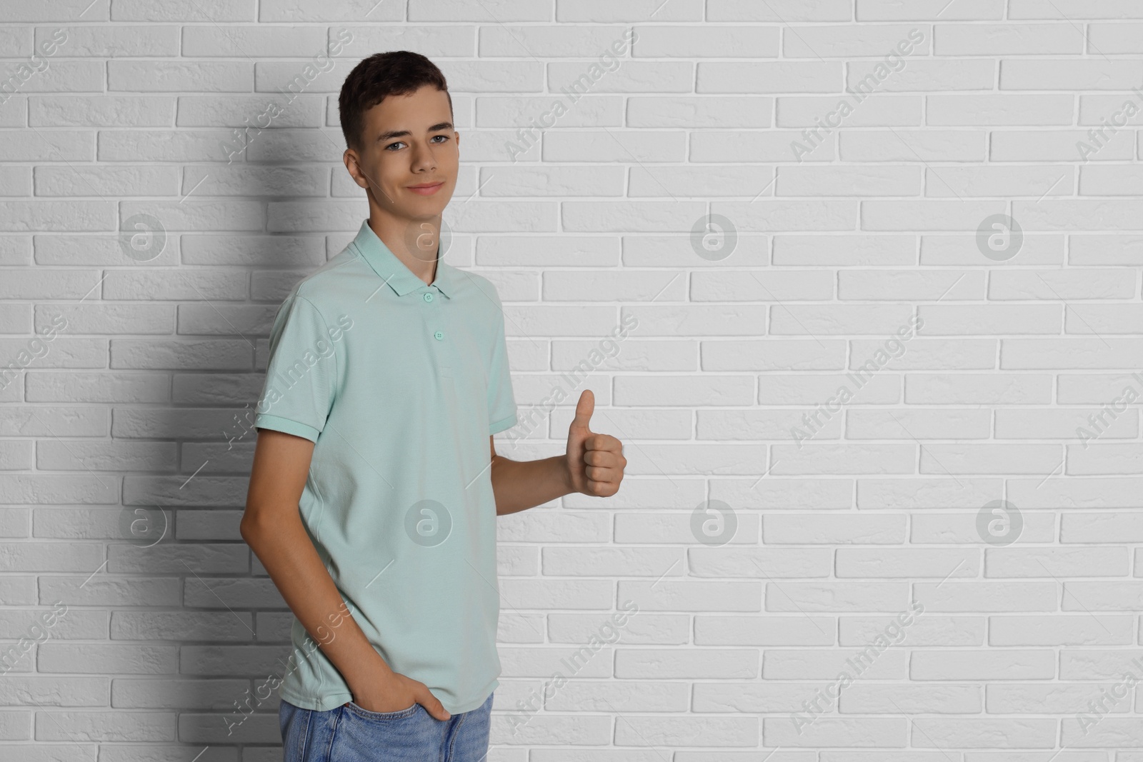 Photo of Teenage boy wearing light green t-shirt and showing thumbs up near white brick wall, space for text