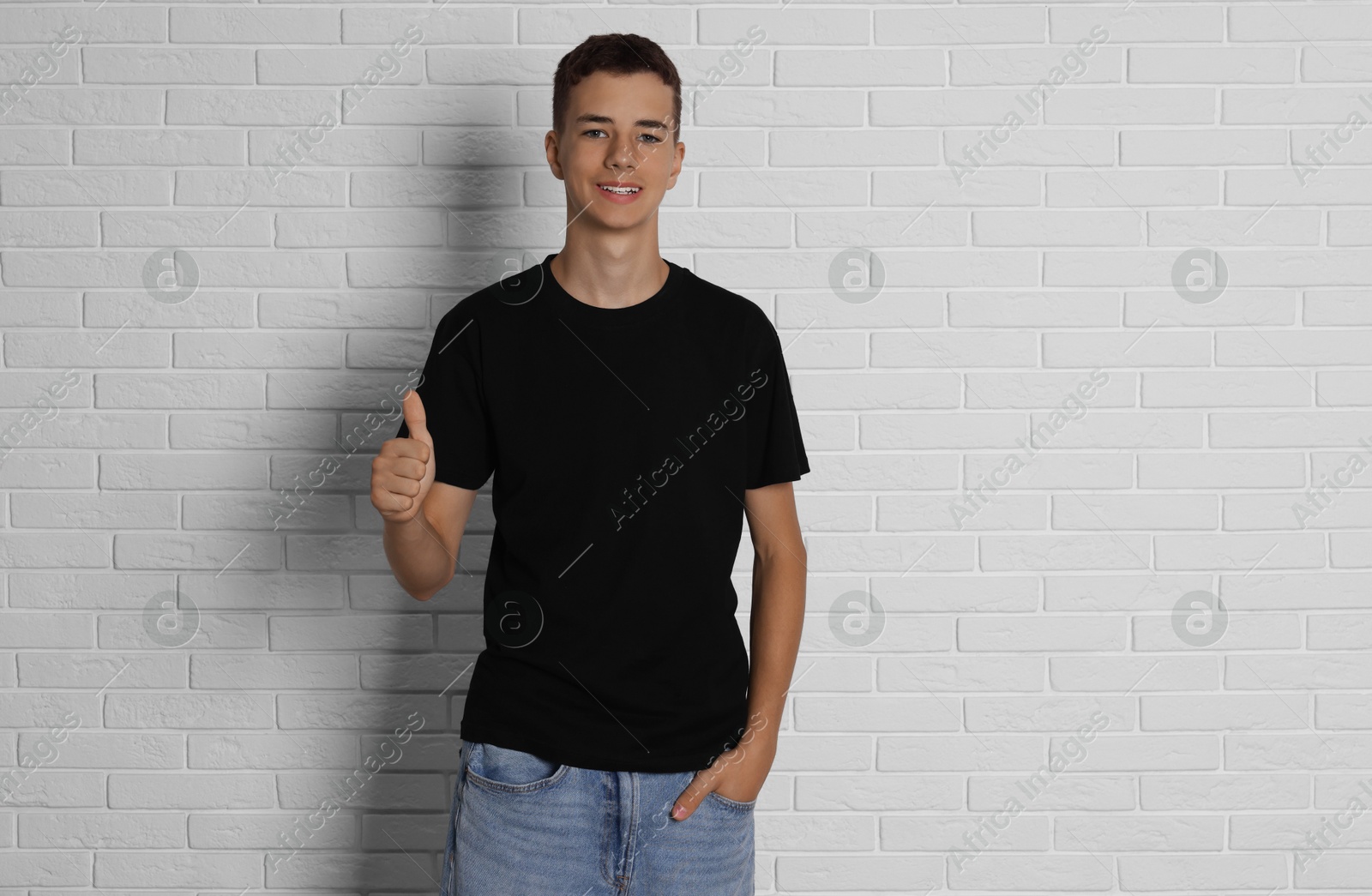 Photo of Teenage boy wearing black t-shirt and showing thumbs up near white brick wall, space for text