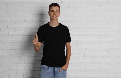 Photo of Teenage boy wearing black t-shirt and showing thumbs up near white brick wall, space for text