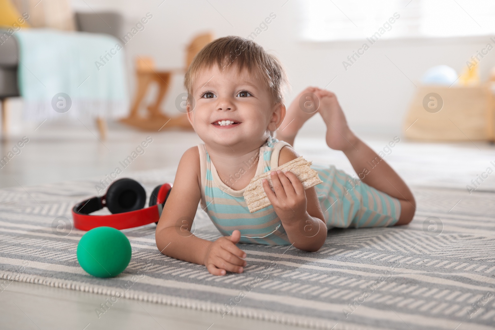 Photo of Cute little baby with snack, ball and headphones on floor at home