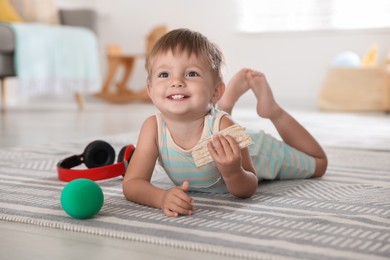 Photo of Cute little baby with snack, ball and headphones on floor at home