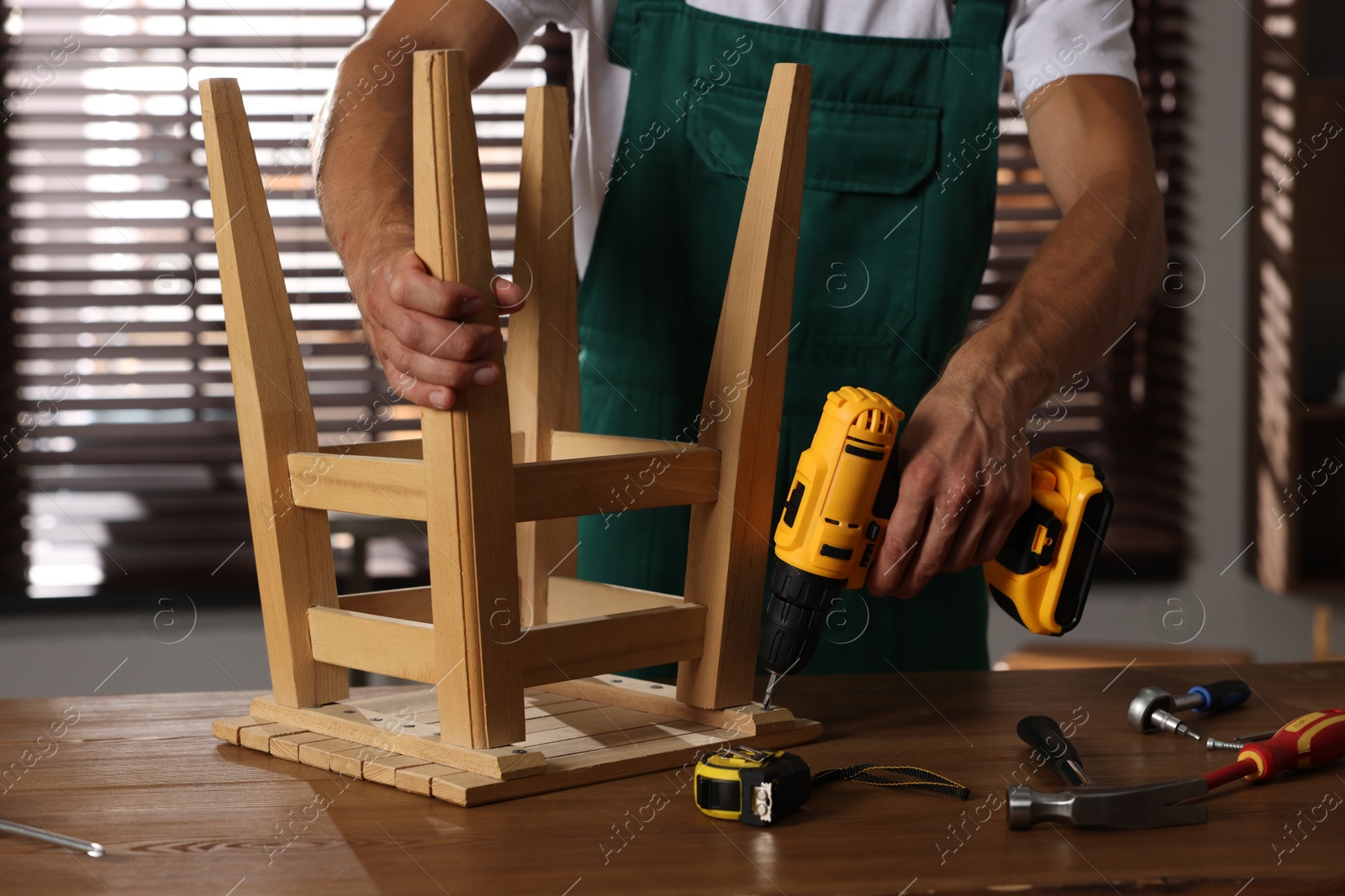 Photo of Man repairing wooden stool with electric screwdriver indoors, closeup