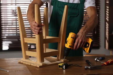 Man repairing wooden stool with electric screwdriver indoors, closeup