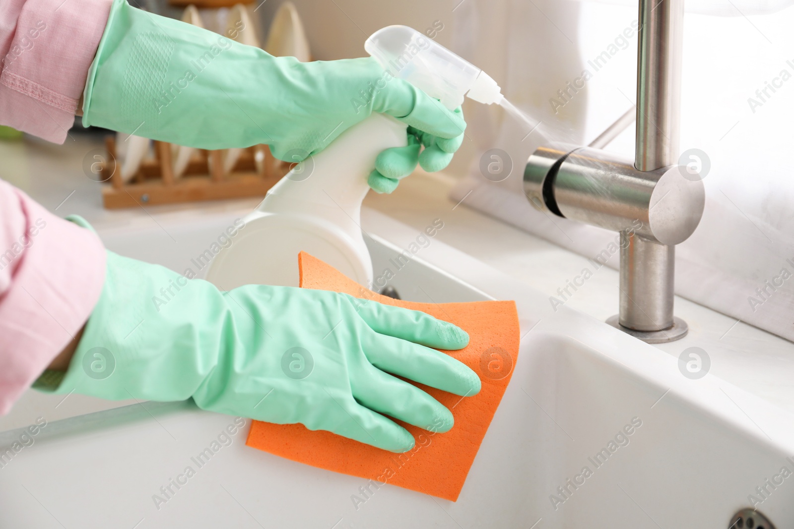 Photo of Woman with spray bottle of detergent and napkin cleaning tap in kitchen, closeup