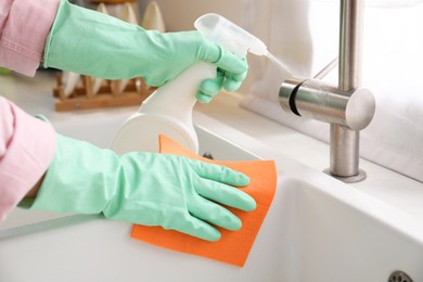 Photo of Woman with spray bottle of detergent and napkin cleaning tap in kitchen, closeup