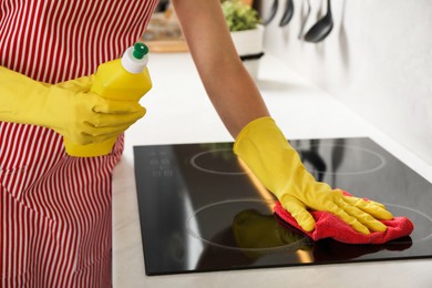 Photo of Woman wiping stove with rag and bottle of detergent in kitchen, closeup