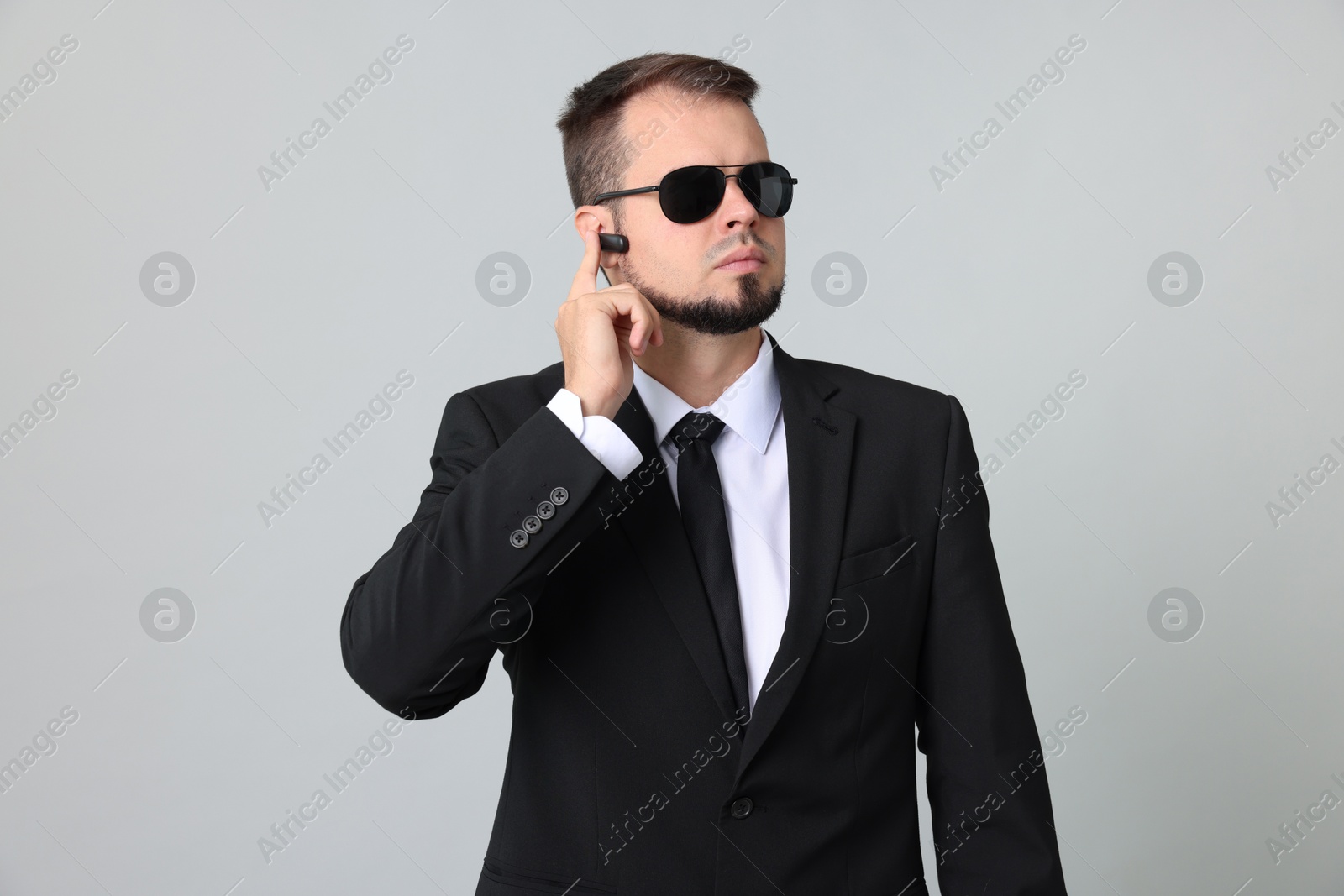 Photo of Young bodyguard in suit and sunglasses using earpiece on grey background