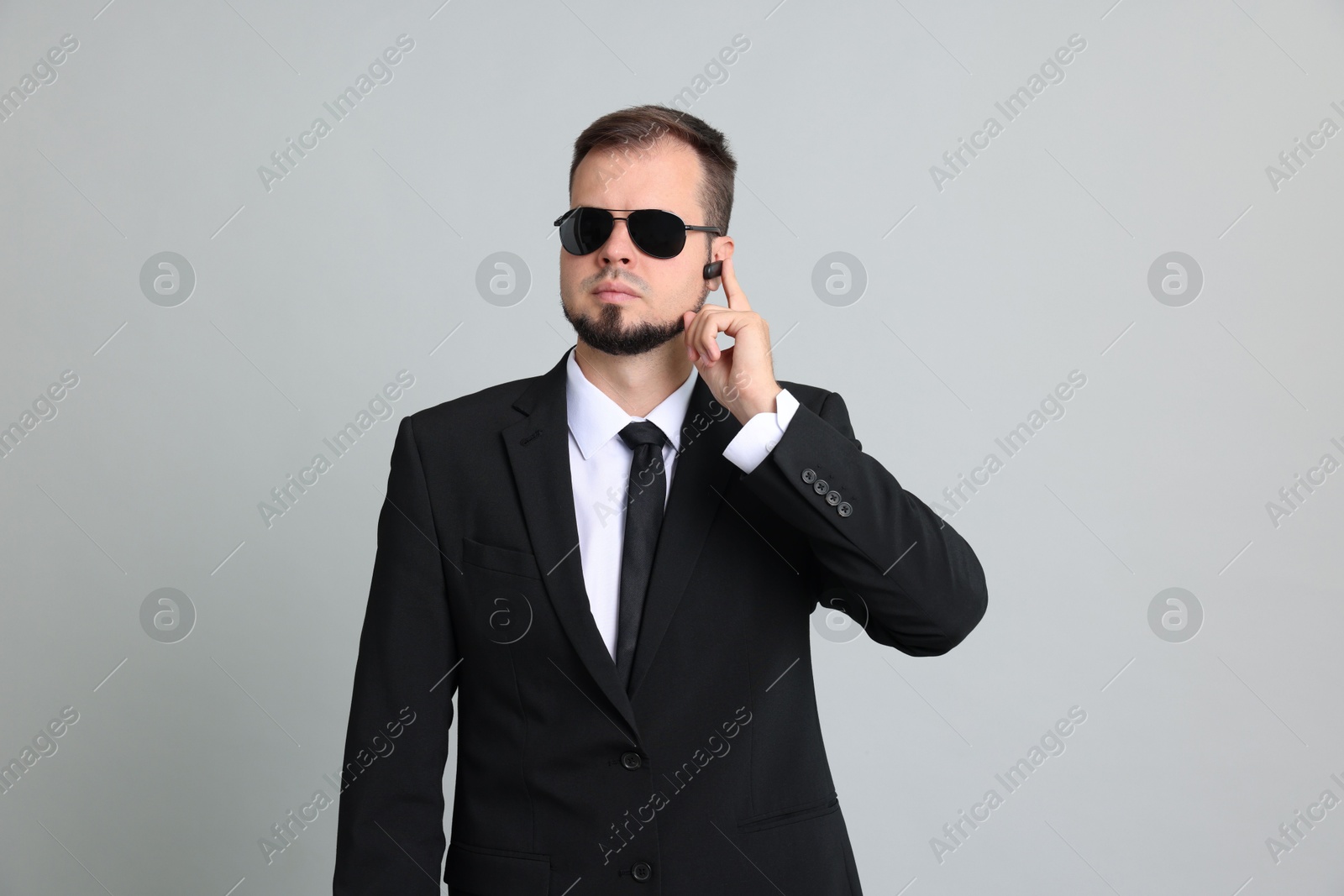 Photo of Young bodyguard in suit and sunglasses using earpiece on grey background