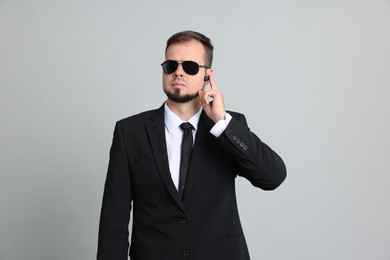 Photo of Young bodyguard in suit and sunglasses using earpiece on grey background