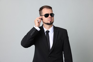 Young bodyguard in suit and sunglasses using earpiece on grey background