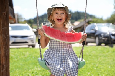 Photo of Cute little girl with slice of juicy watermelon on swing outdoors