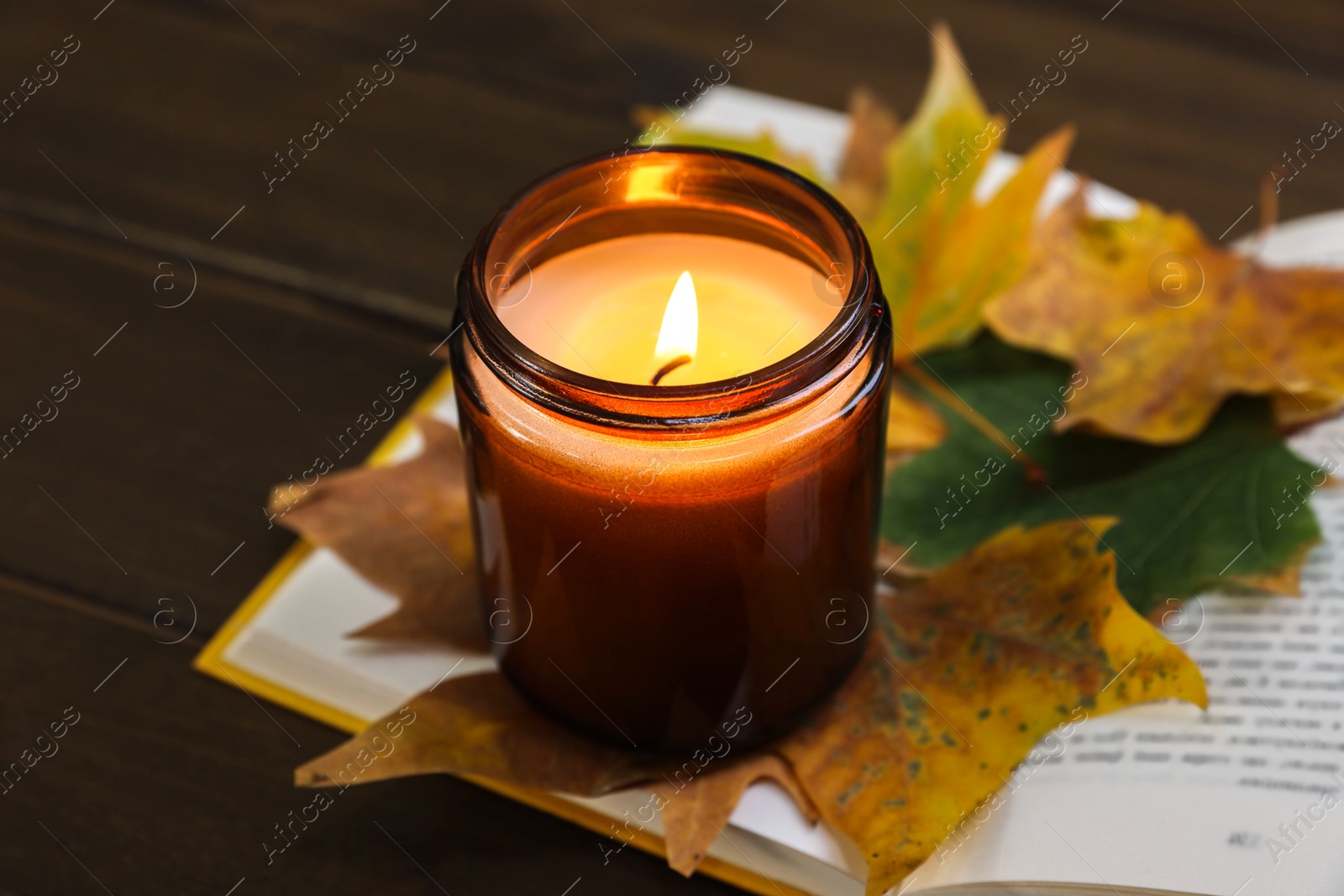 Photo of Burning candle, dry leaves and open book on wooden table, closeup. Autumn atmosphere