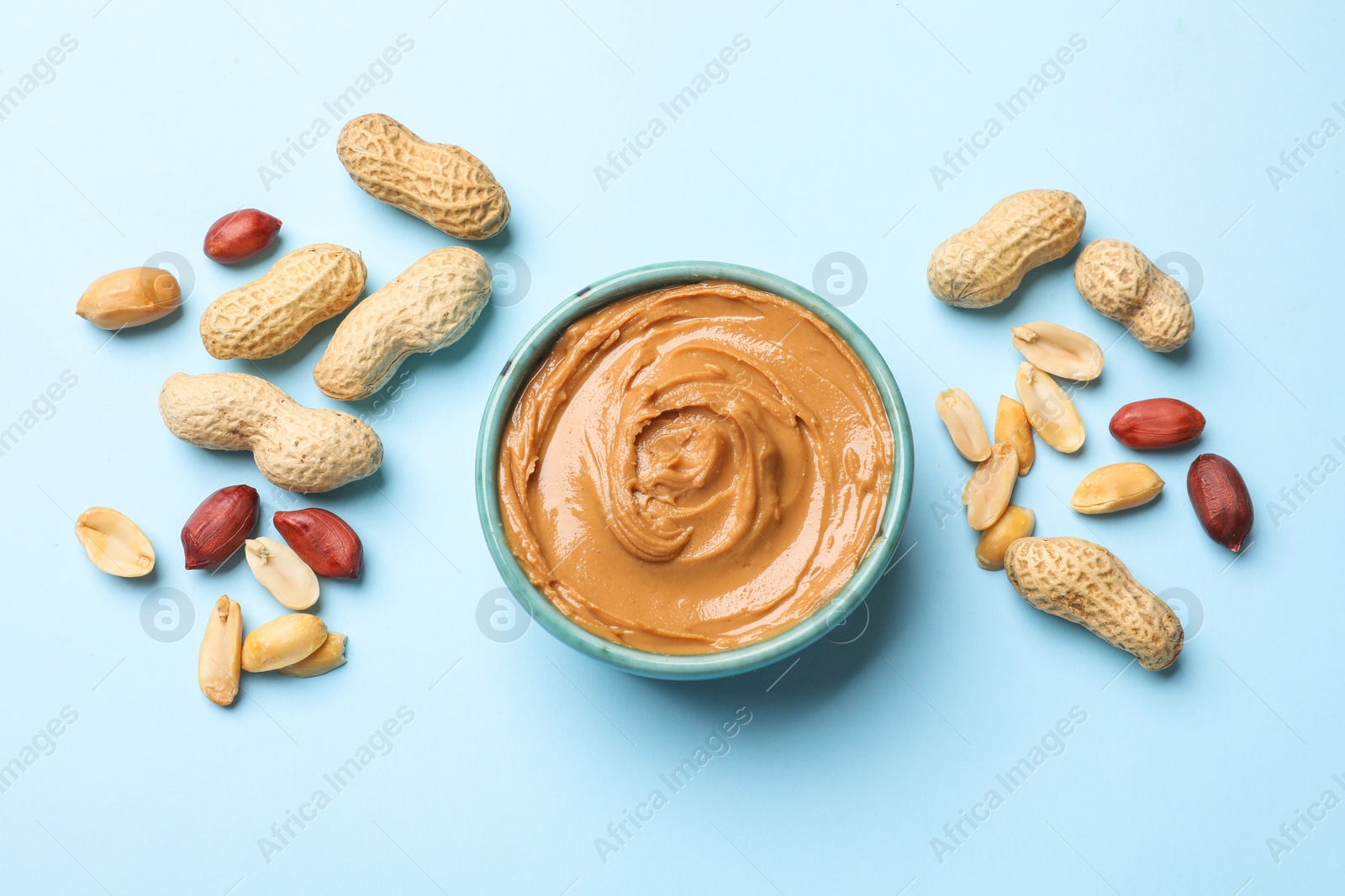 Photo of Tasty peanut butter in bowl and groundnuts on light blue background, flat lay