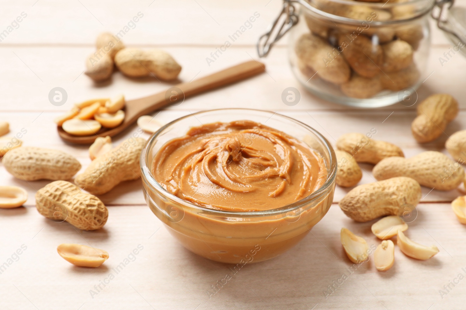 Photo of Tasty peanut butter in bowl, groundnuts, jar and spoon on wooden table, closeup