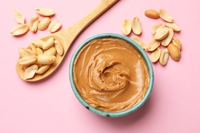 Photo of Tasty peanut butter in bowl and spoon with groundnuts on pink table, flat lay