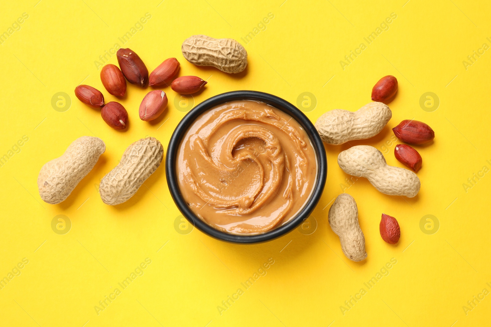 Photo of Tasty peanut butter in bowl and groundnuts on yellow table, flat lay