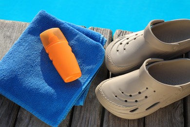 Photo of Stylish beach slippers, towel and sunscreen on wooden deck near outdoor swimming pool, above view