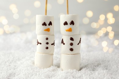 Photo of Cute snowmen made of marshmallows on artificial snow against blurred lights