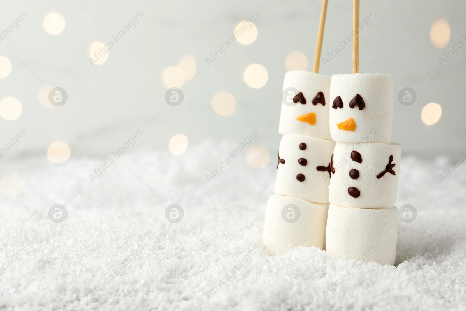 Photo of Cute snowmen made of marshmallows on artificial snow against blurred lights. Space for text