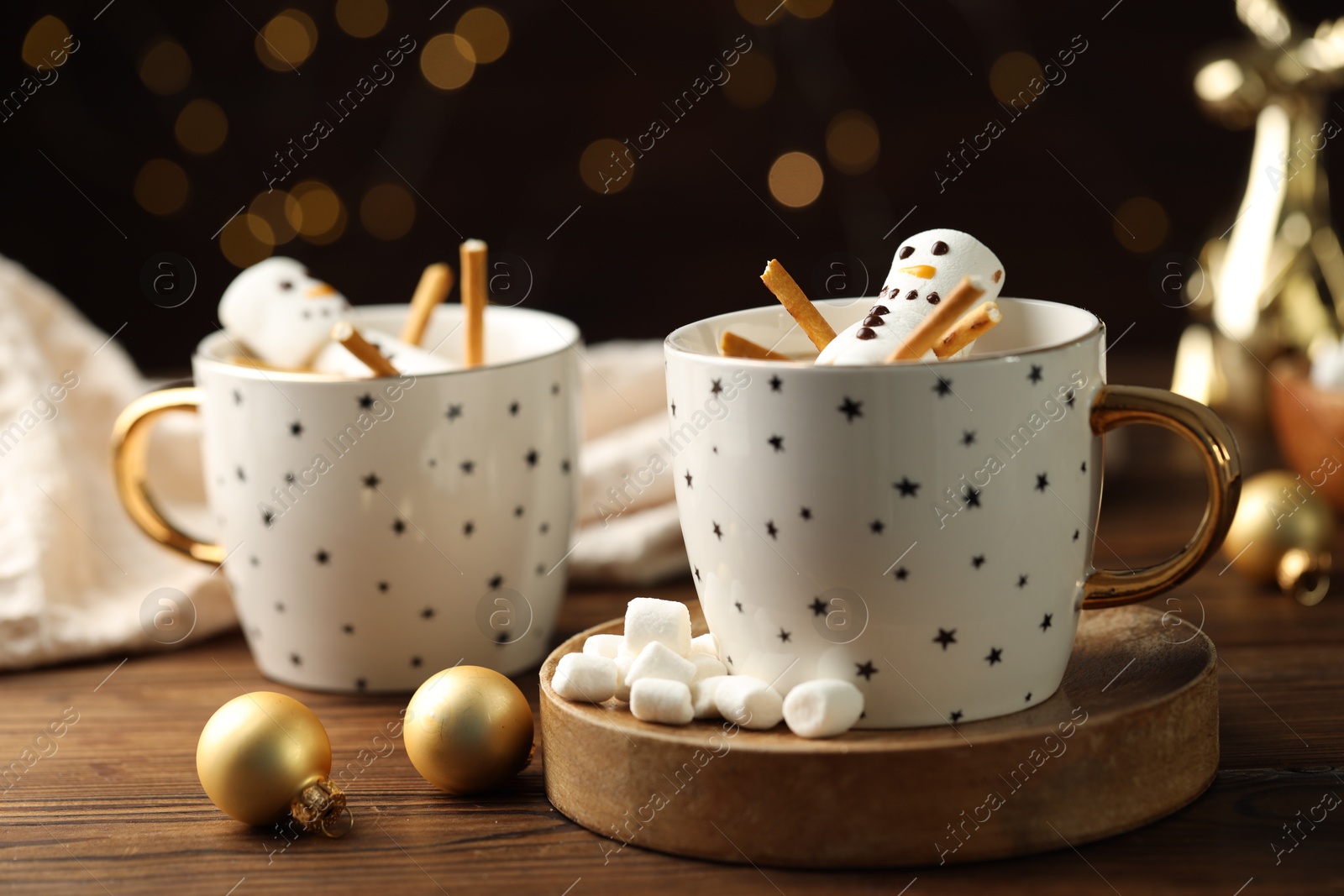 Photo of Cute marshmallow snowmen in cups of hot drinks on wooden table with Christmas decor against blurred lights, closeup