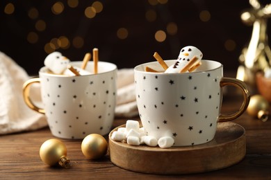 Photo of Cute marshmallow snowmen in cups of hot drinks on wooden table with Christmas decor against blurred lights, closeup