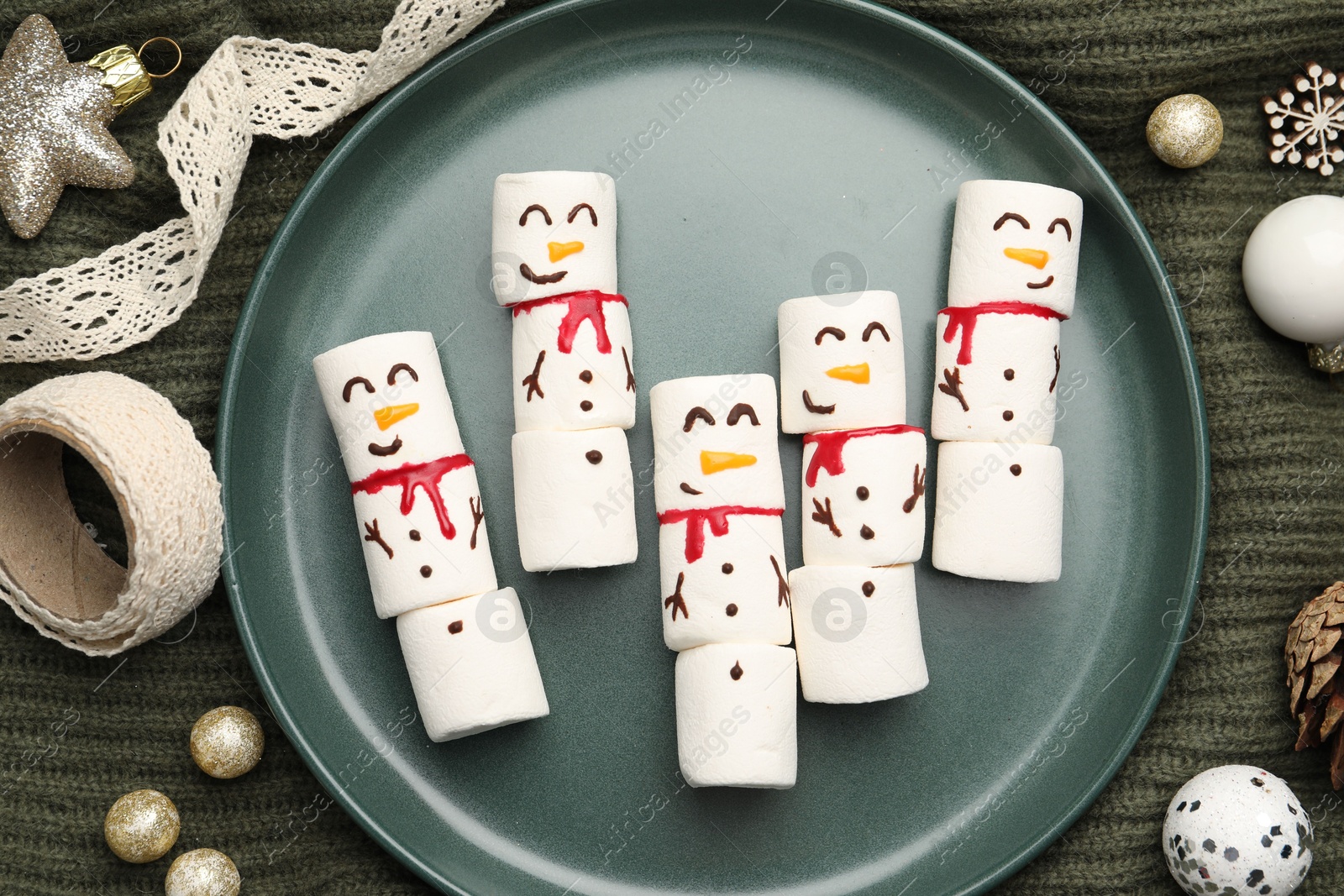 Photo of Funny marshmallow snowmen and festive decor on olive knitted fabric, top view