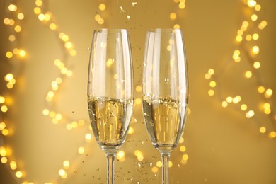 Photo of Glasses of sparkling wine and confetti against golden background with blurred lights, closeup. Christmas decor