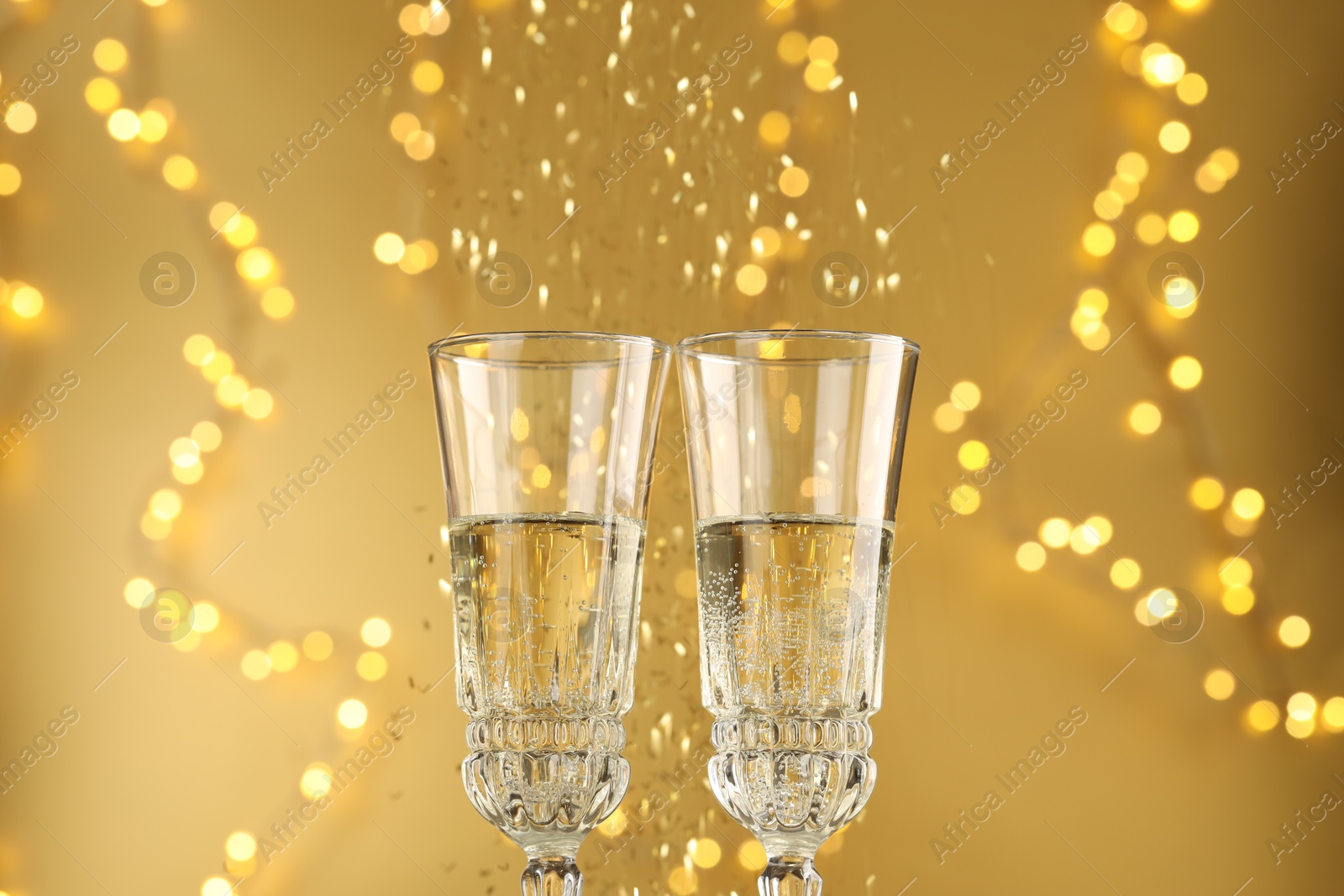 Photo of Glasses of sparkling wine and confetti against golden background with blurred lights, closeup. Christmas decor