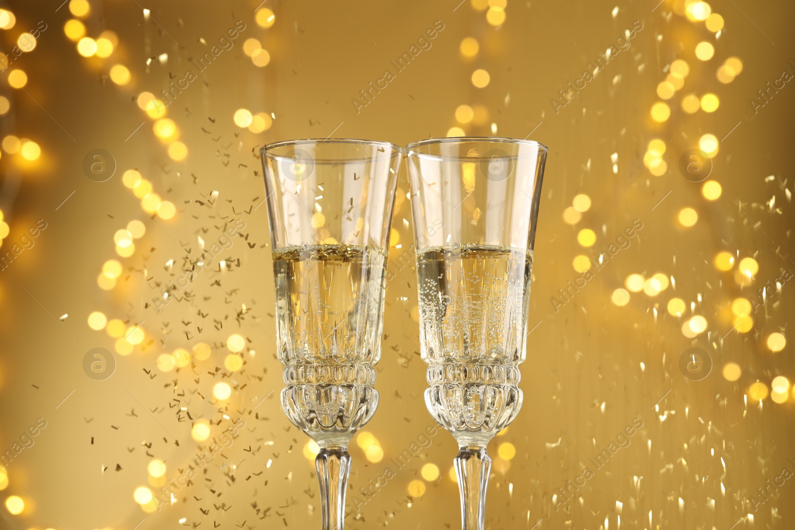 Photo of Glasses of sparkling wine and confetti against golden background with blurred lights, closeup. Christmas decor