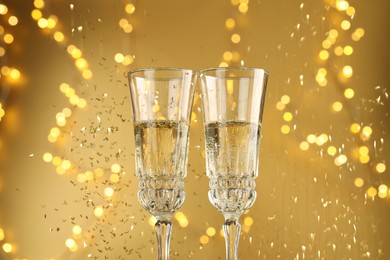 Photo of Glasses of sparkling wine and confetti against golden background with blurred lights, closeup. Christmas decor