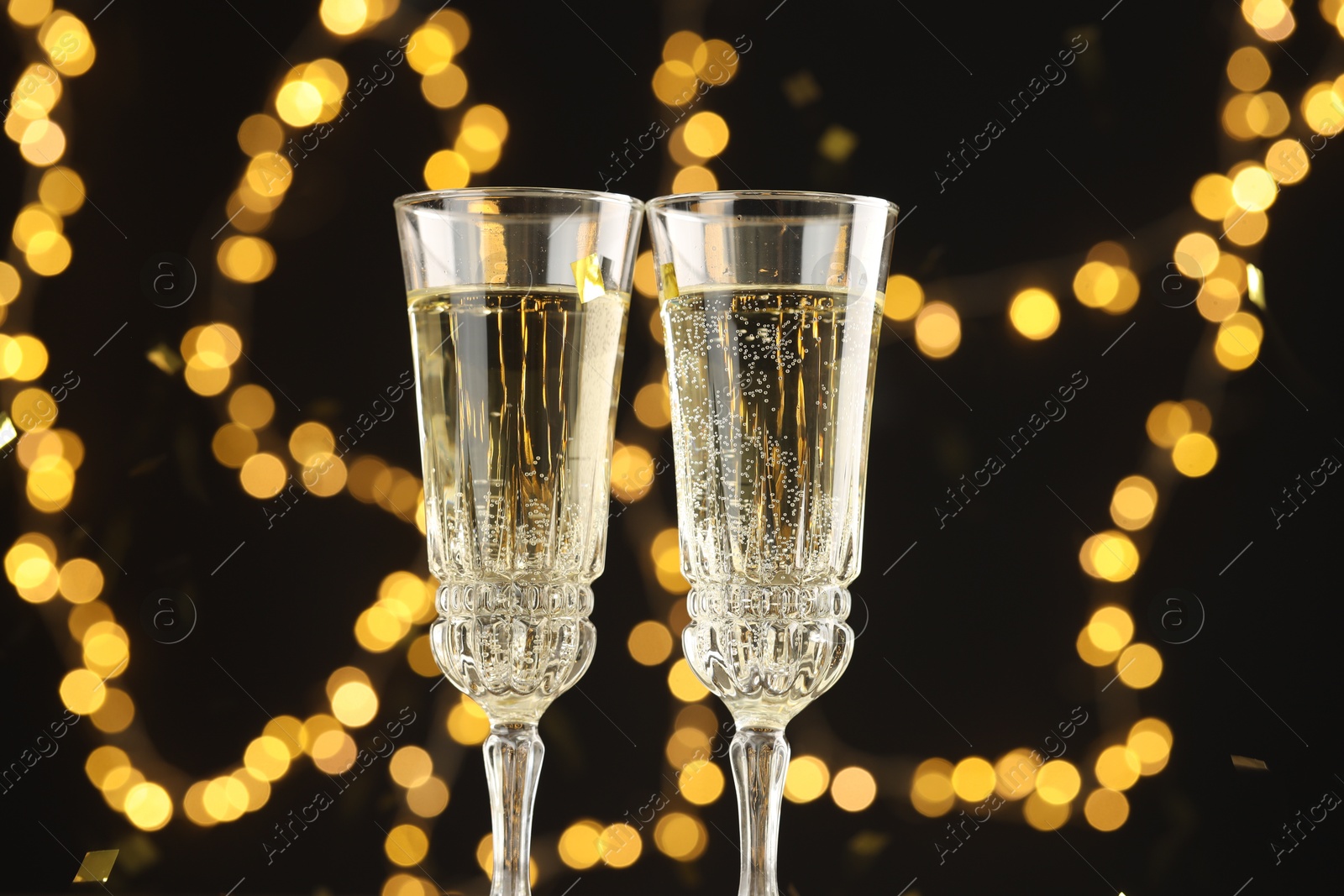 Photo of Glasses of sparkling wine and confetti against dark background with blurred lights, closeup. Christmas decor