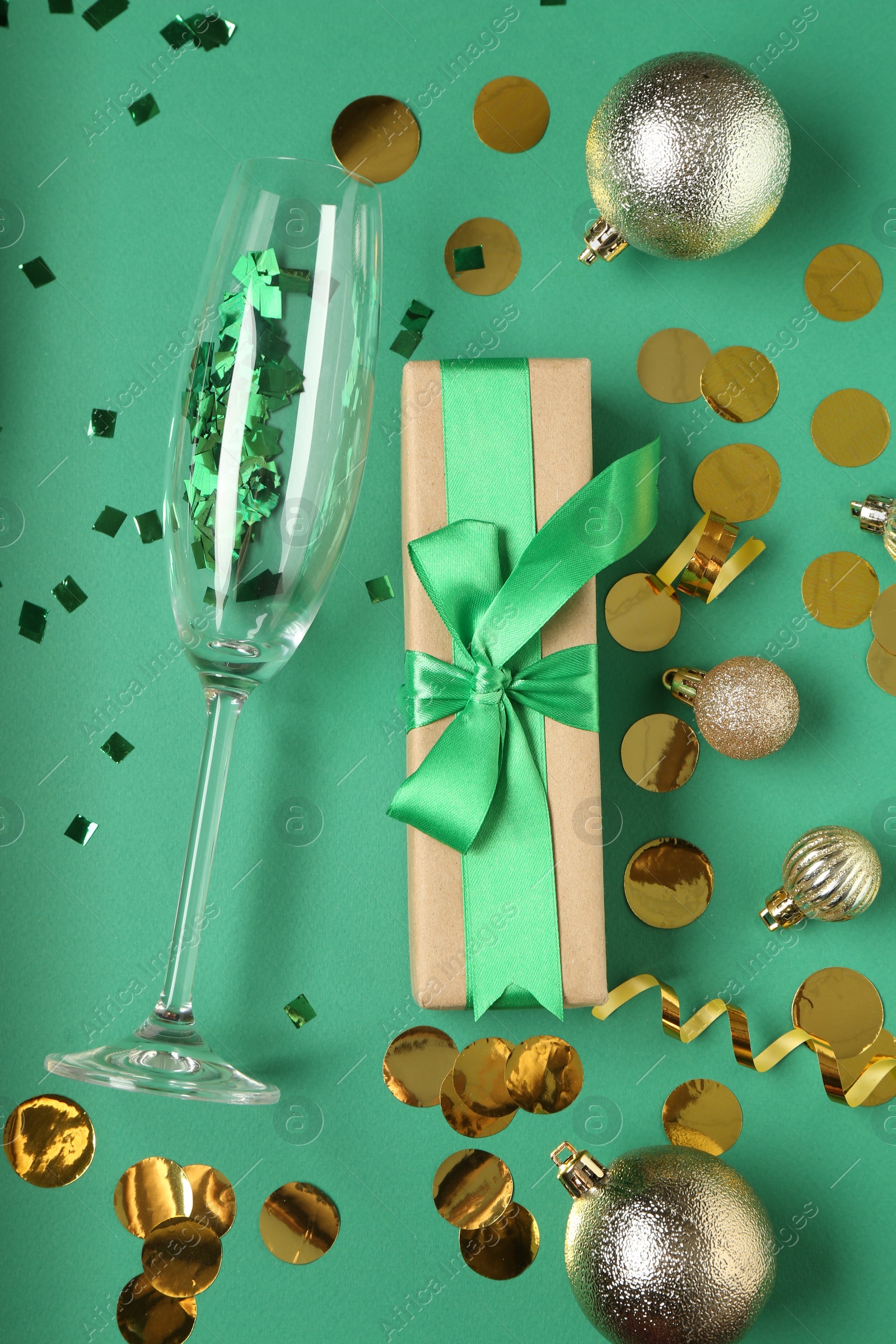 Photo of Glass with confetti, gift box and Christmas balls on green background, flat lay