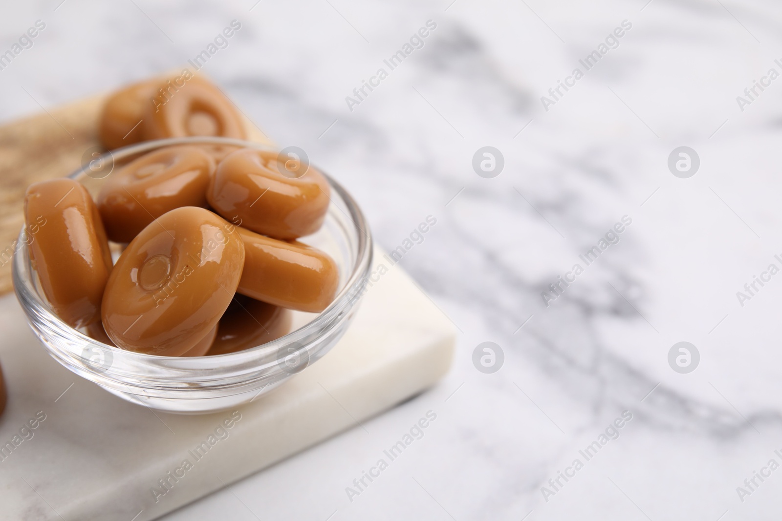 Photo of Tasty hard toffee candies in bowl on white marble table, closeup. Space for text