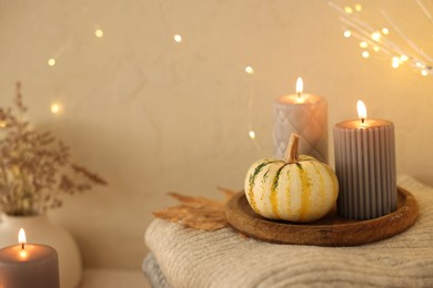 Photo of Burning candles, pumpkins and stack of sweaters near beige wall, space for text. Autumn atmosphere