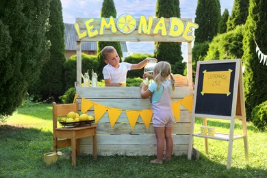 Cute boy standing at lemonade stand while little girl pouring refreshing drink into paper cup in park