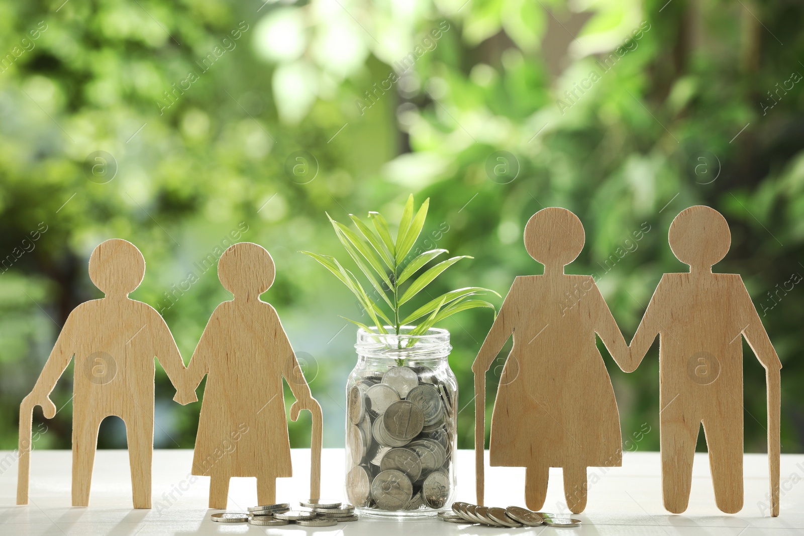 Photo of Pension savings. Figures of elderly people, jar with coins and twig on white table against blurred green background