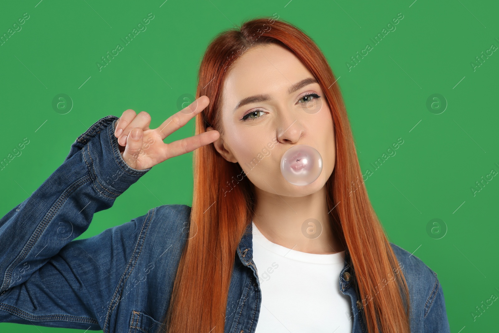 Photo of Beautiful woman blowing bubble gum and showing peace gesture on green background