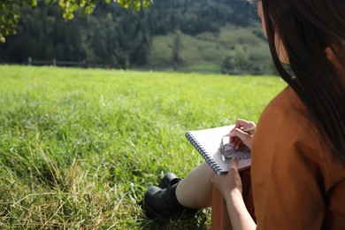 Photo of Beautiful young woman drawing with pencil in notepad outdoors on green grass, closeup