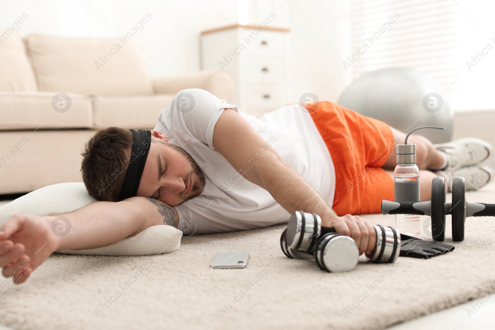 Photo of Lazy young man with sport equipment sleeping on floor at home