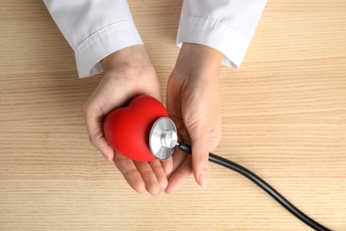 Photo of Doctor holding red heart and stethoscope on wooden background, top view. Cardiology concept