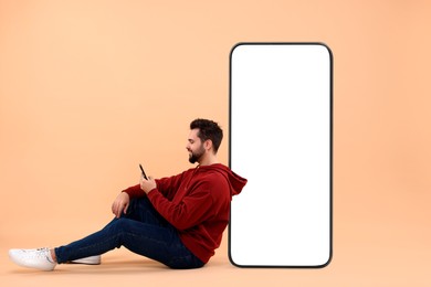 Image of Man with mobile phone sitting near huge device with empty screen on dark beige background. Mockup for design