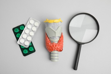Plastic model of thyroid with tumor, pills and magnifying glass on grey background, flat lay